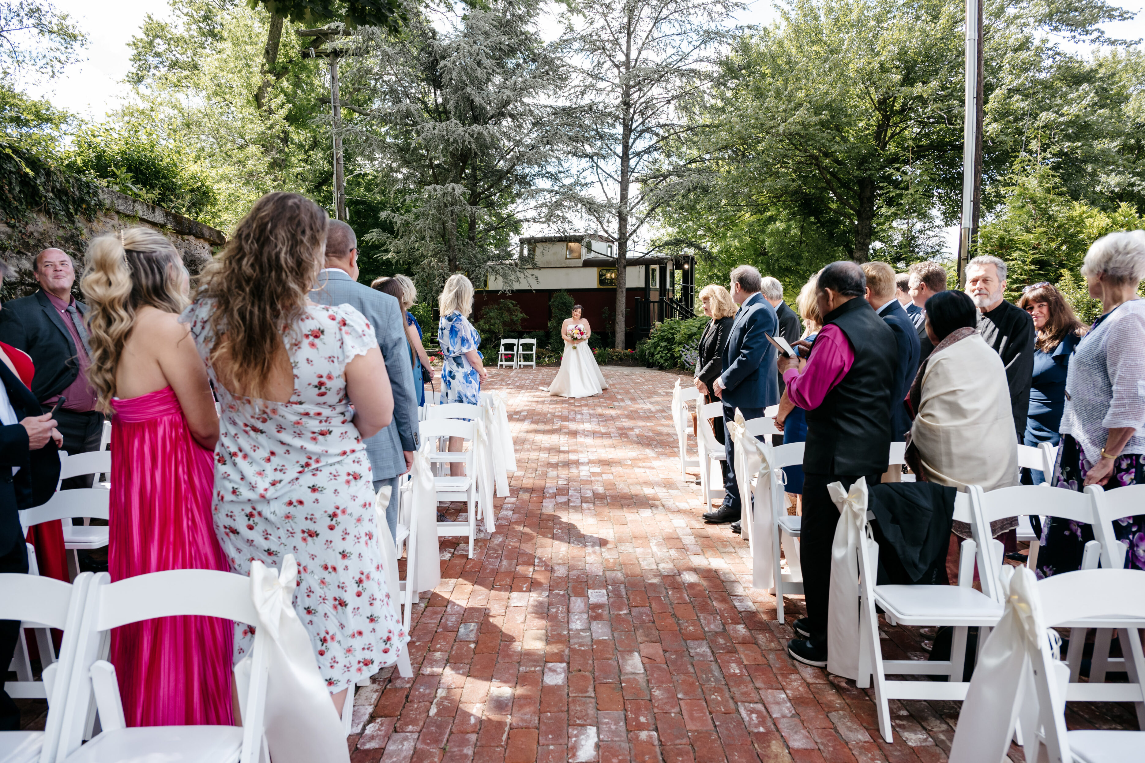 Bride walks alone down a brick aisle outdoors as guests stand on either side under trees and sunlight.