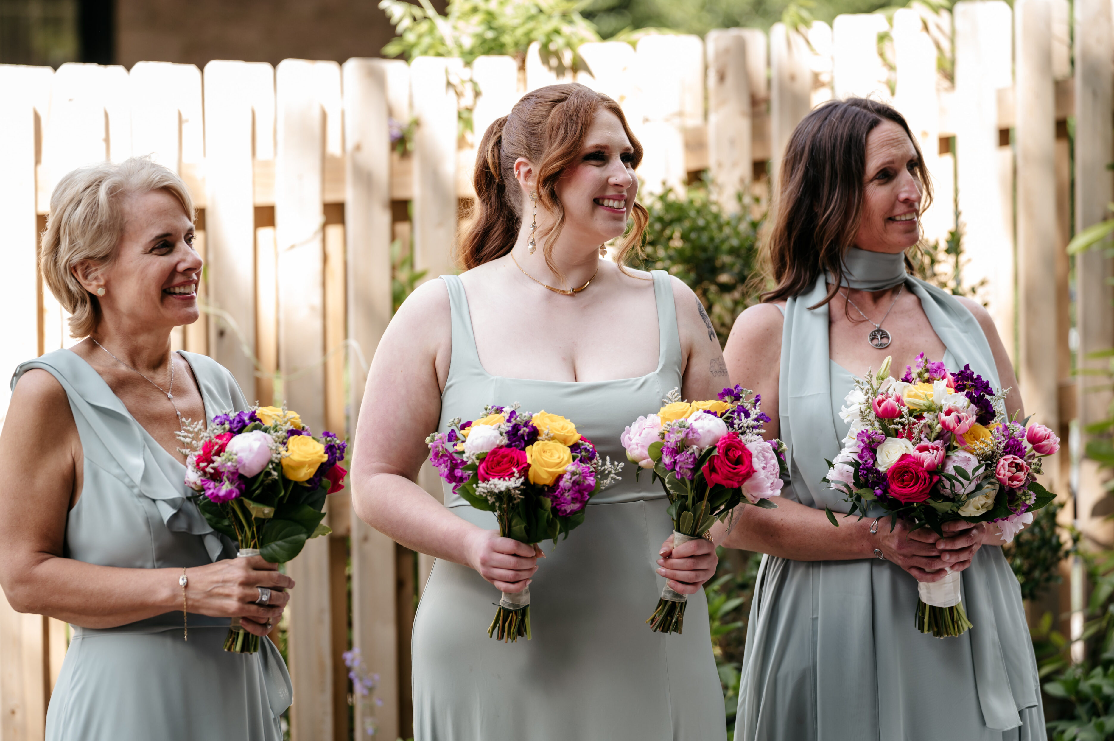 Three women in light green dresses hold colorful bouquets, smiling outdoors in front of a wooden fence.