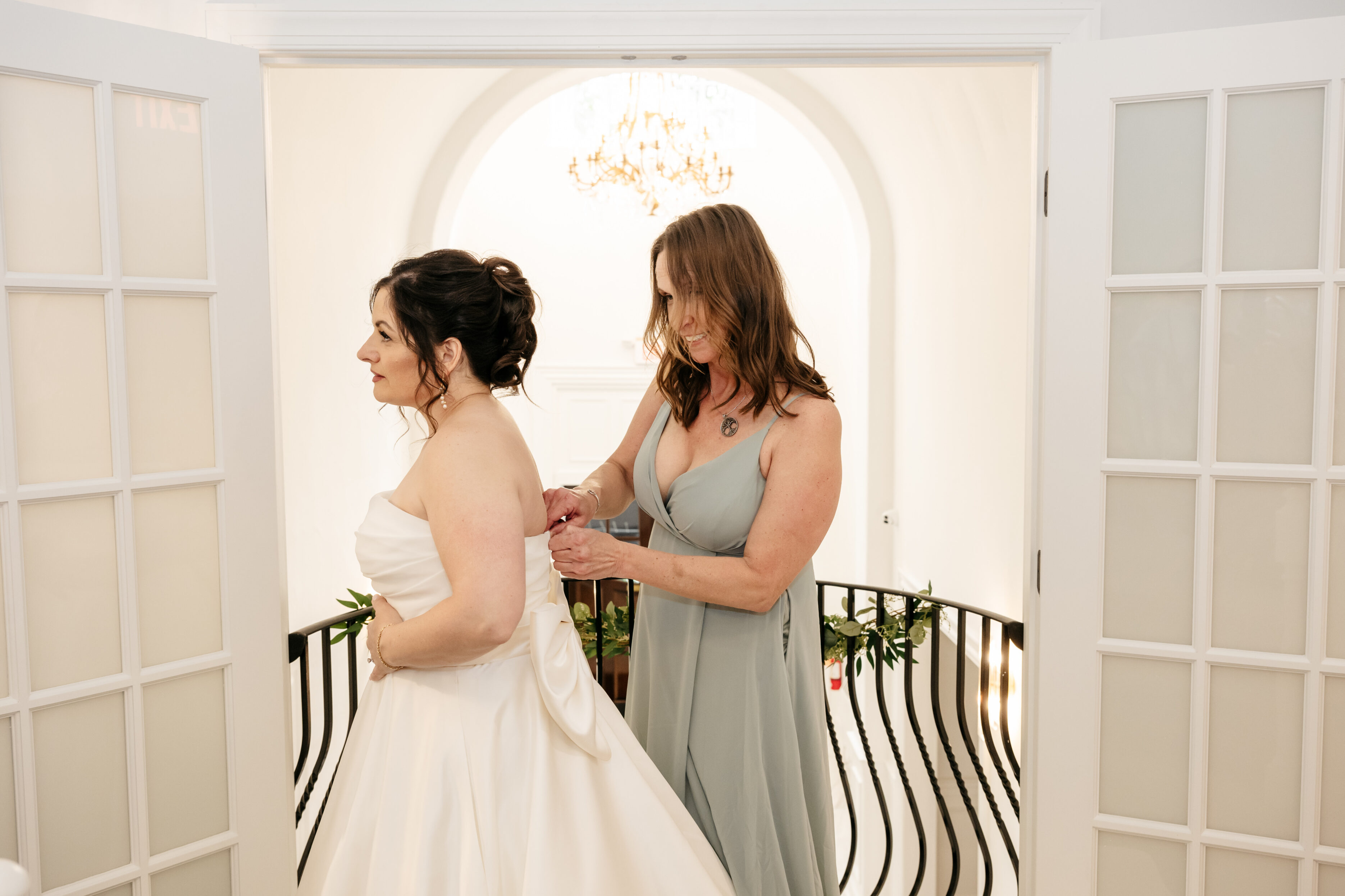 Woman in a blue dress helps a bride adjust her white gown in a bright, elegant room with double doors.