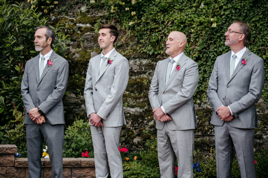 Four men in light gray suits with boutonnieres stand outside in front of a stone wall and greenery.