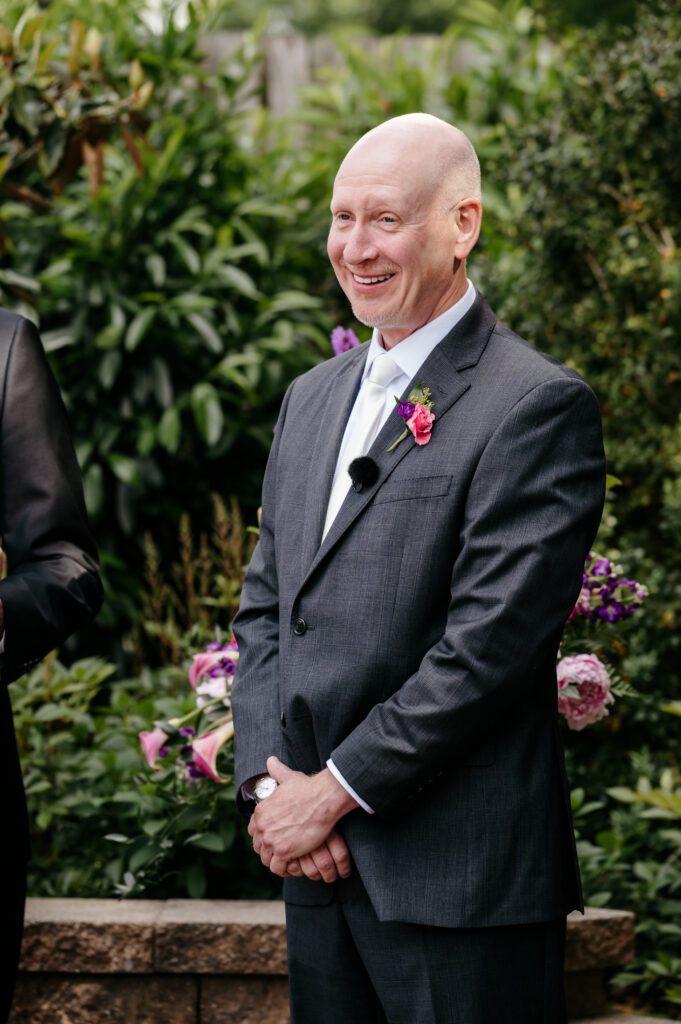 A man in a suit smiles outside, standing with hands clasped in front of greenery and purple flowers.