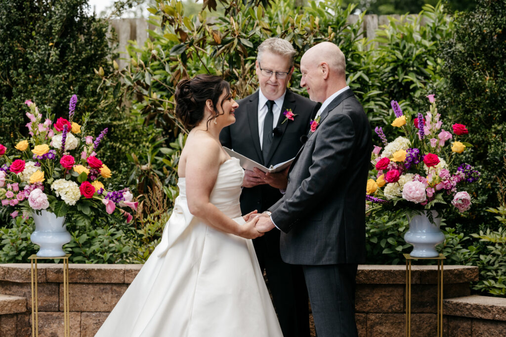 A bride and groom hold hands and smile at each other during an outdoor wedding ceremony with a celebrant and flowers.