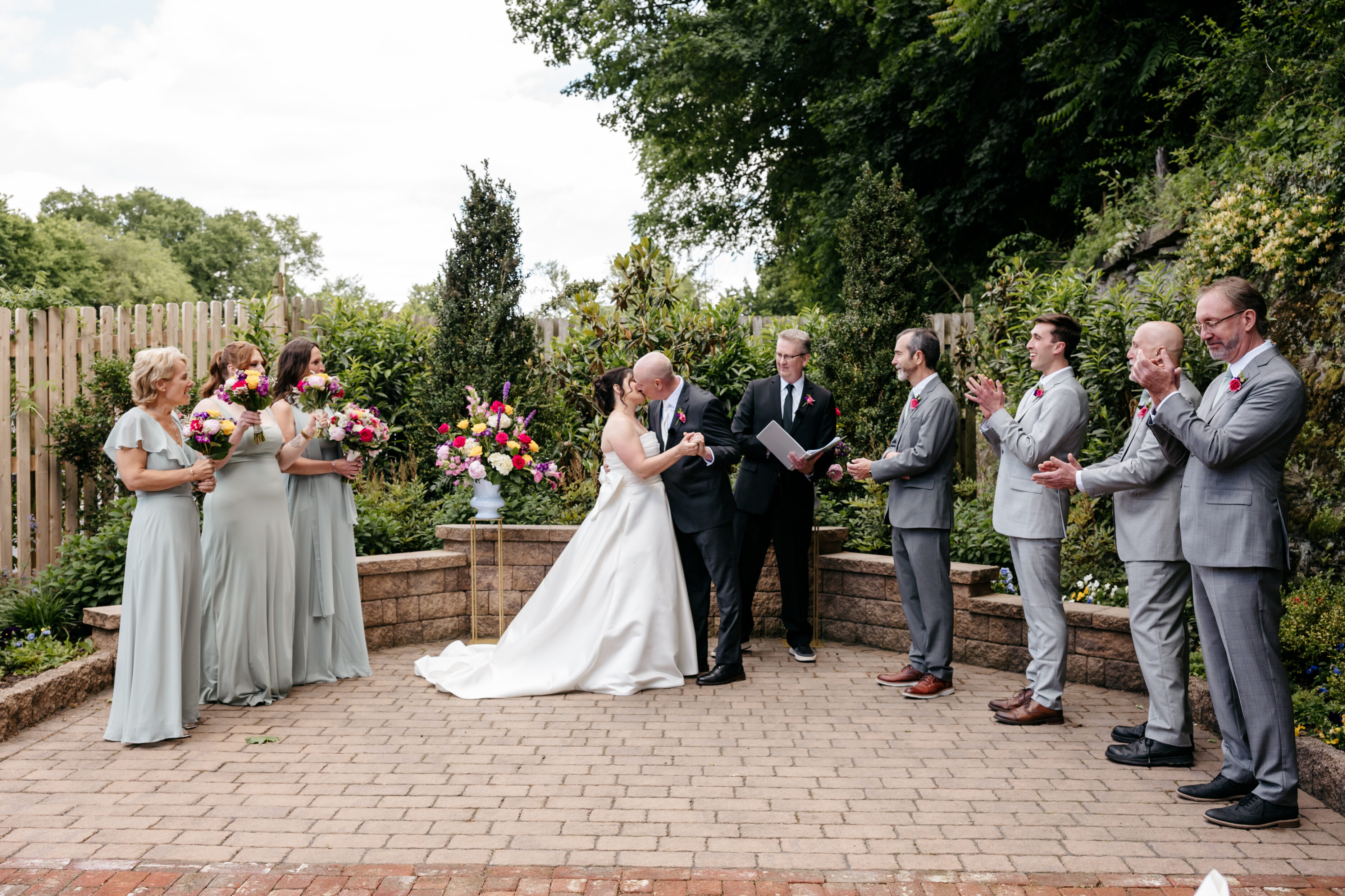 A bride and groom kiss at their outdoor wedding ceremony, surrounded by bridesmaids and groomsmen.