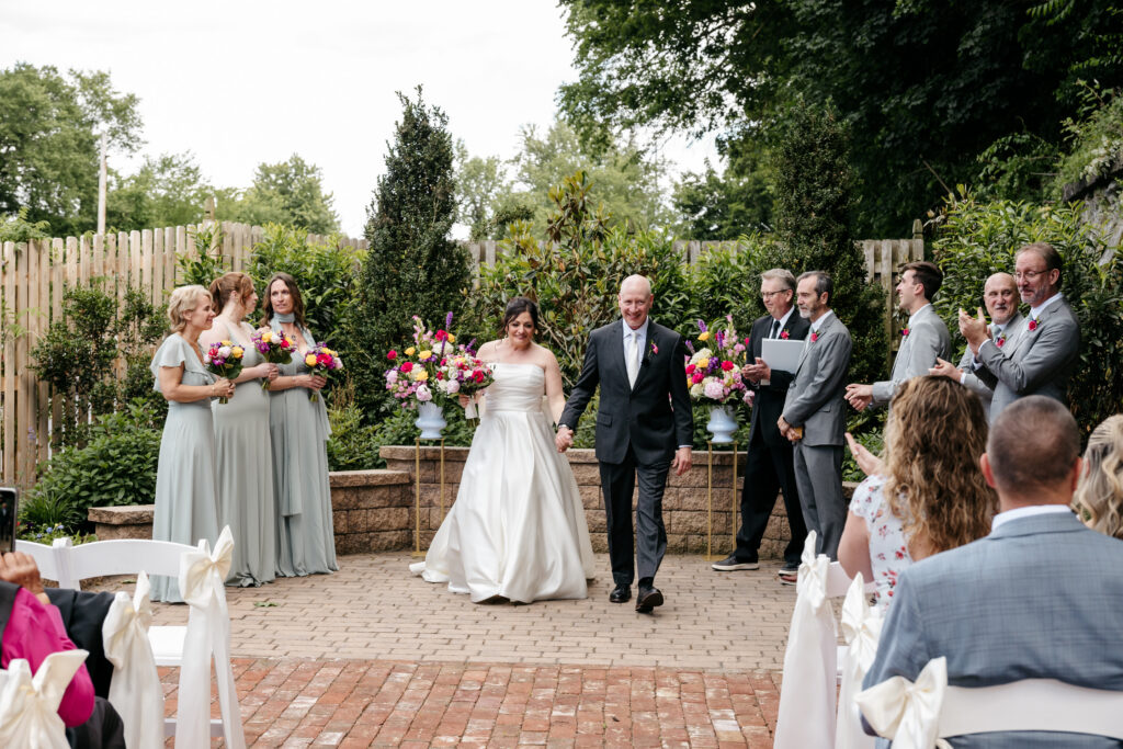 A bride in a white dress walks down the aisle outdoors, escorted by an older man, as guests look on.