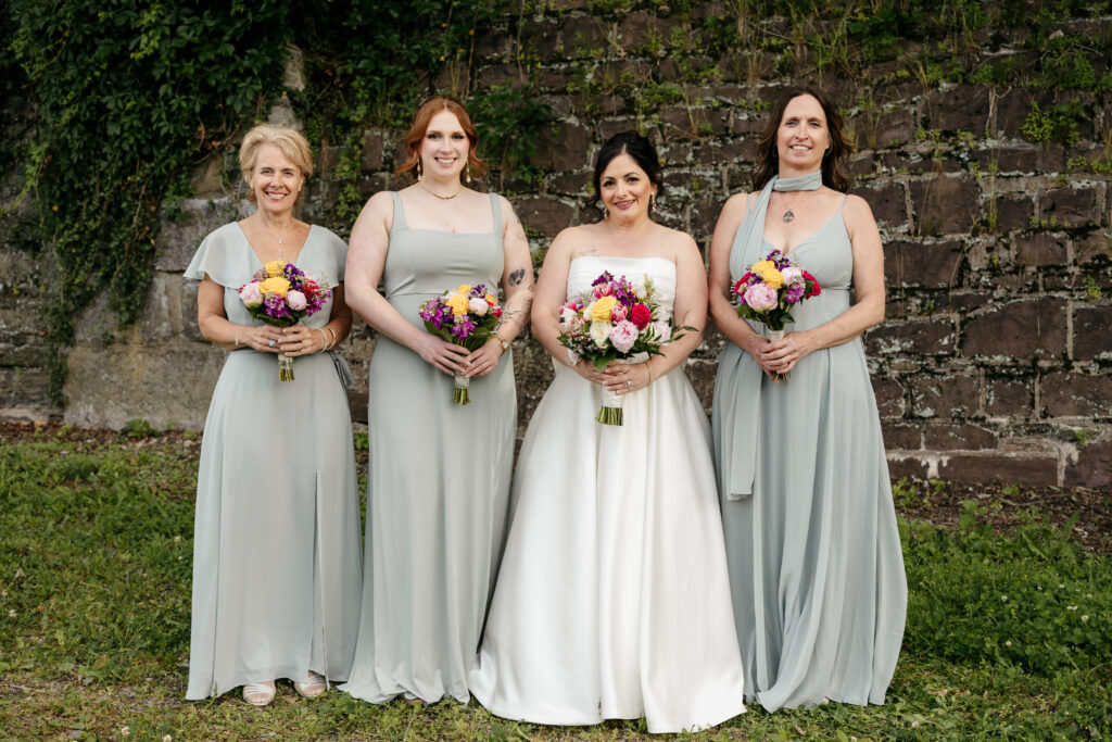 Four women in formal dresses pose outdoors with bouquets in front of a stone wall covered in greenery.