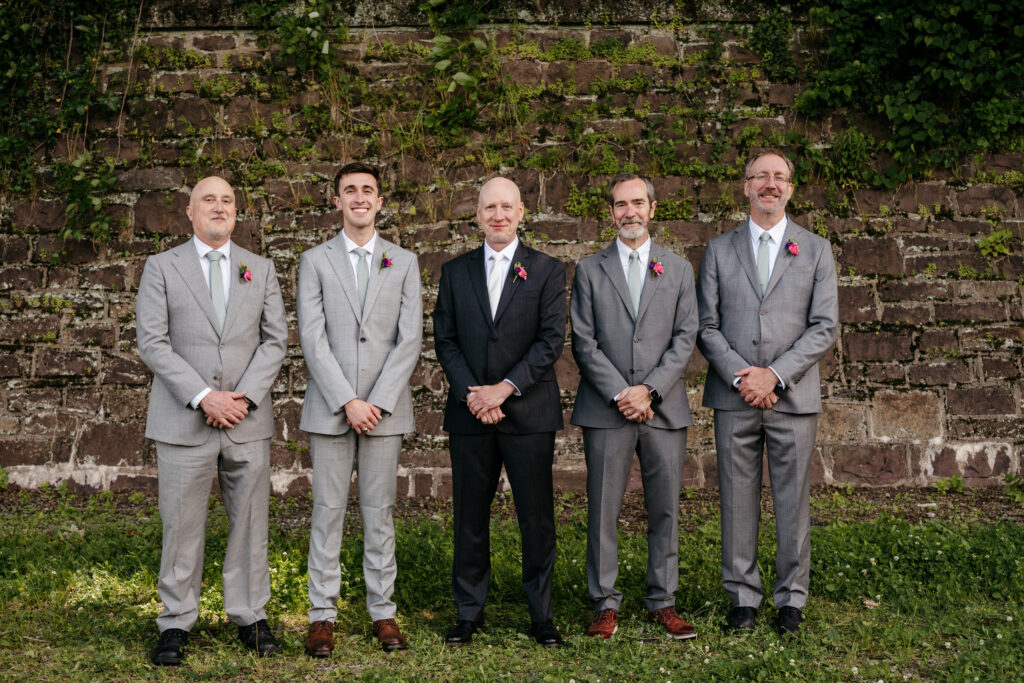 Five men in suits stand side by side in front of a stone wall covered in greenery, smiling at the camera.