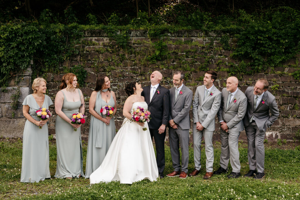 A bride and groom laugh with their wedding party, all smiling, standing outdoors in front of a stone wall.