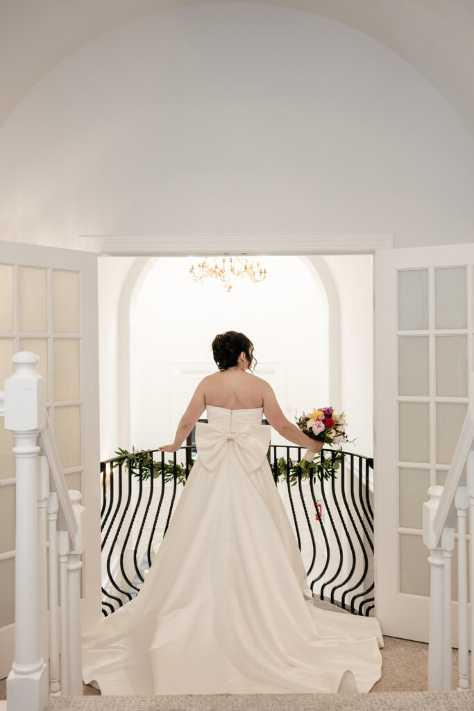 Bride in a white gown with a large bow stands on a balcony holding a bouquet, facing away from the camera.