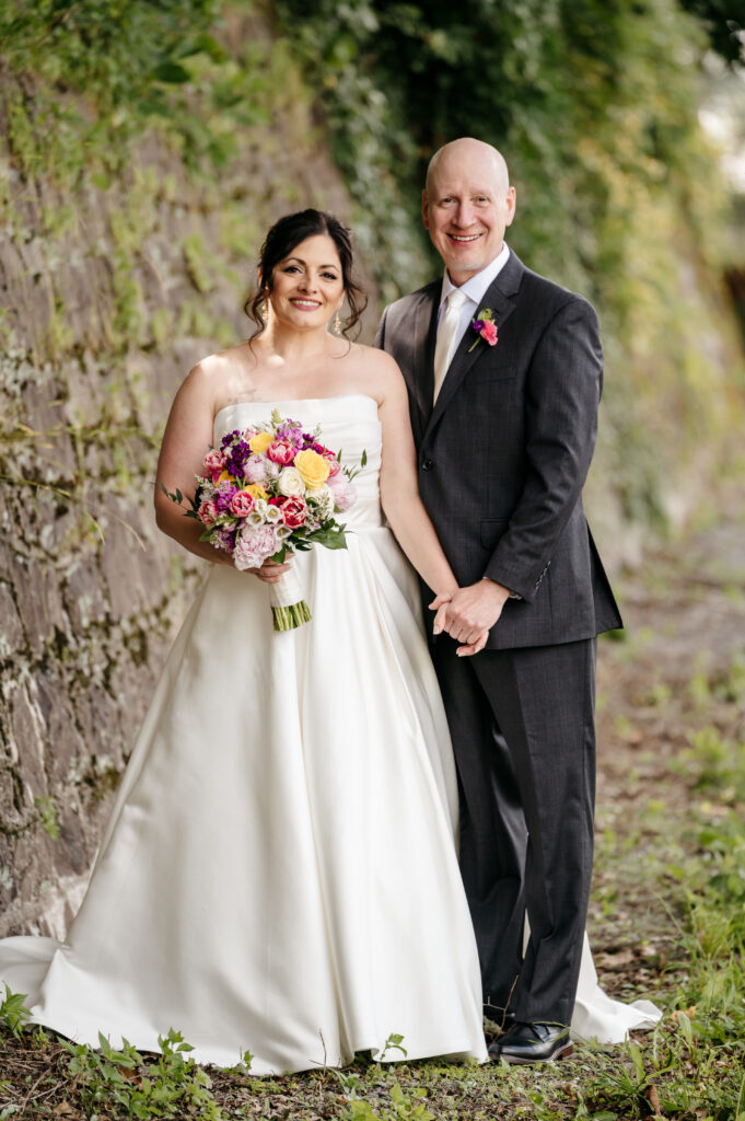 A bride holding a bouquet stands beside a groom in a suit, both smiling outdoors near a stone wall and greenery.
