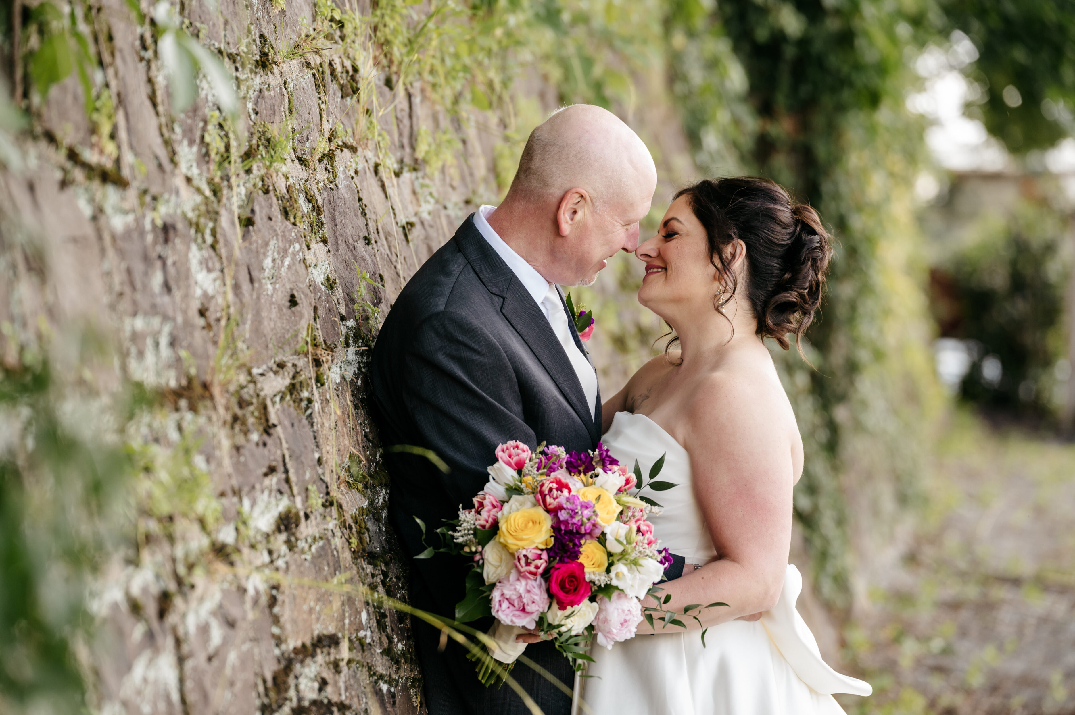 A bride and groom smile at each other, embracing by a stone wall, bride holding a colorful bouquet.
