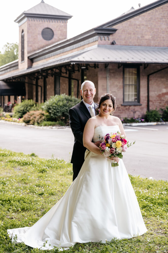 A bride and groom smile outdoors; she holds a bouquet, and they stand in front of a stone building.