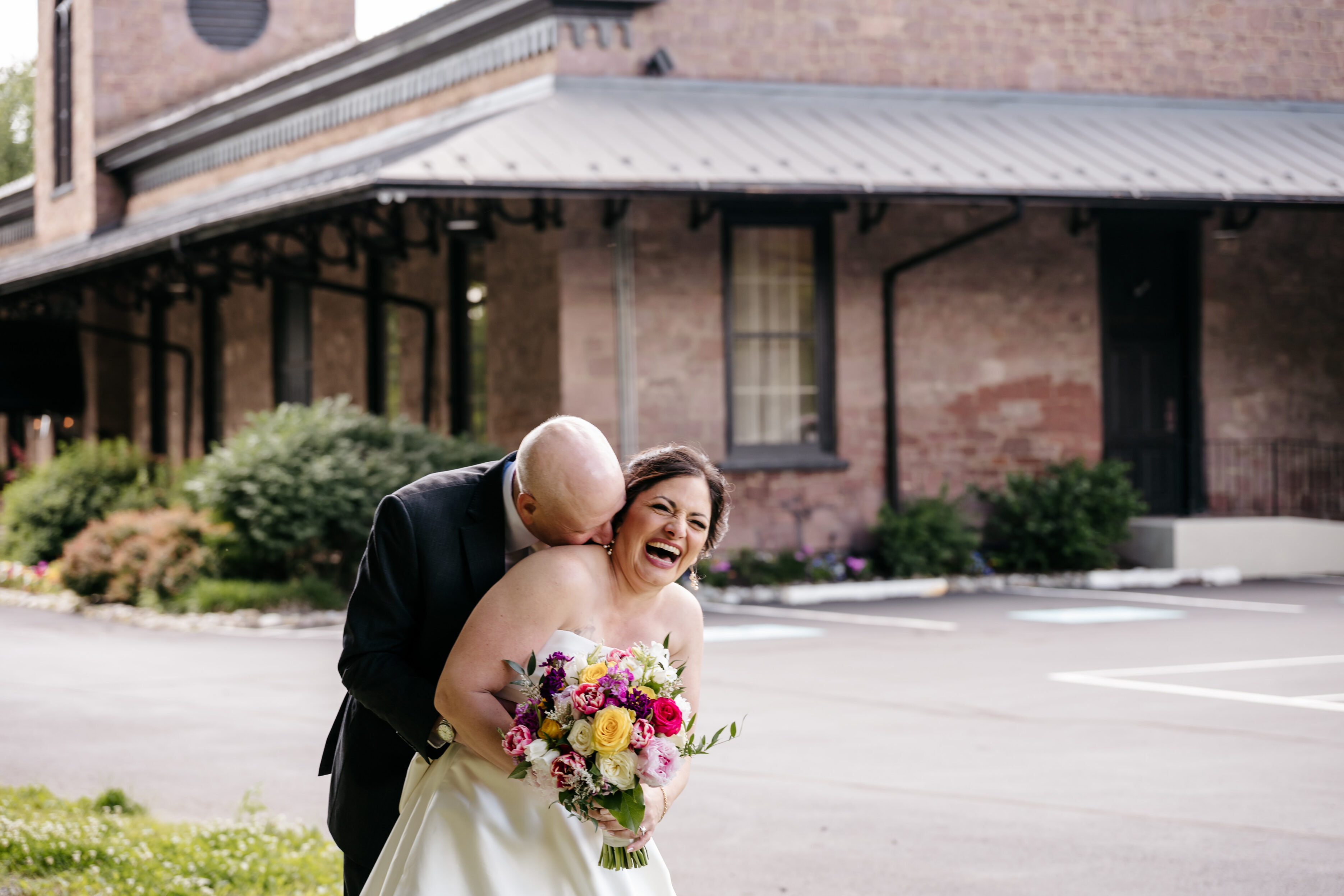 A groom playfully hugs a laughing bride holding a colorful bouquet outside a brick building.