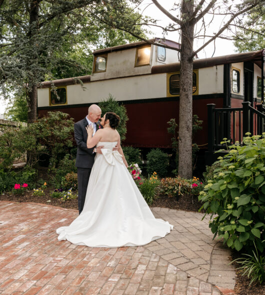 A bride and groom embrace on a brick path in front of a red train car surrounded by greenery and flowers.