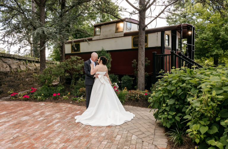 A bride and groom embrace on a brick path in front of a red train car surrounded by greenery and flowers.