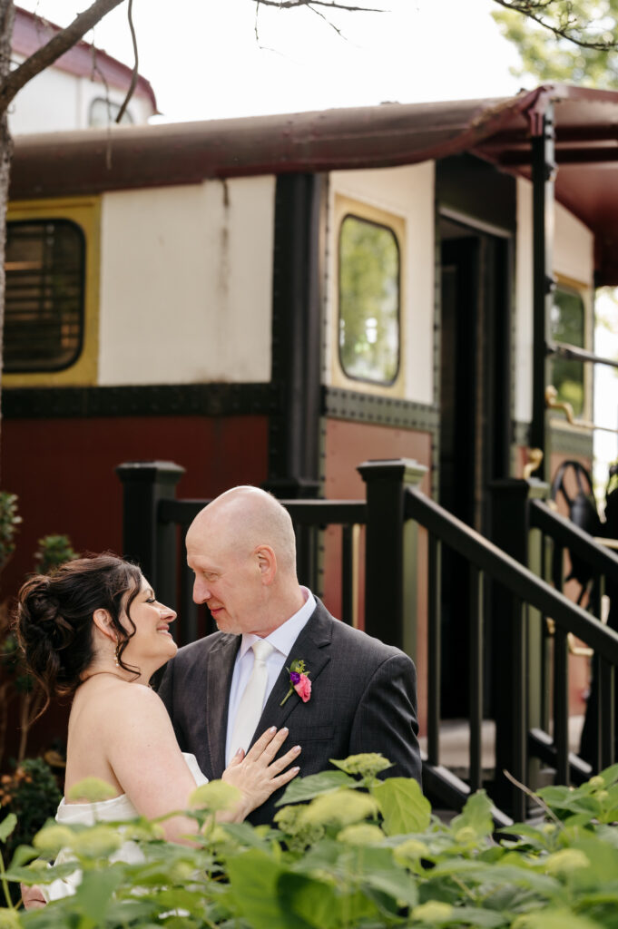 A couple in formal attire smiles at each other in front of a vintage train car surrounded by greenery.