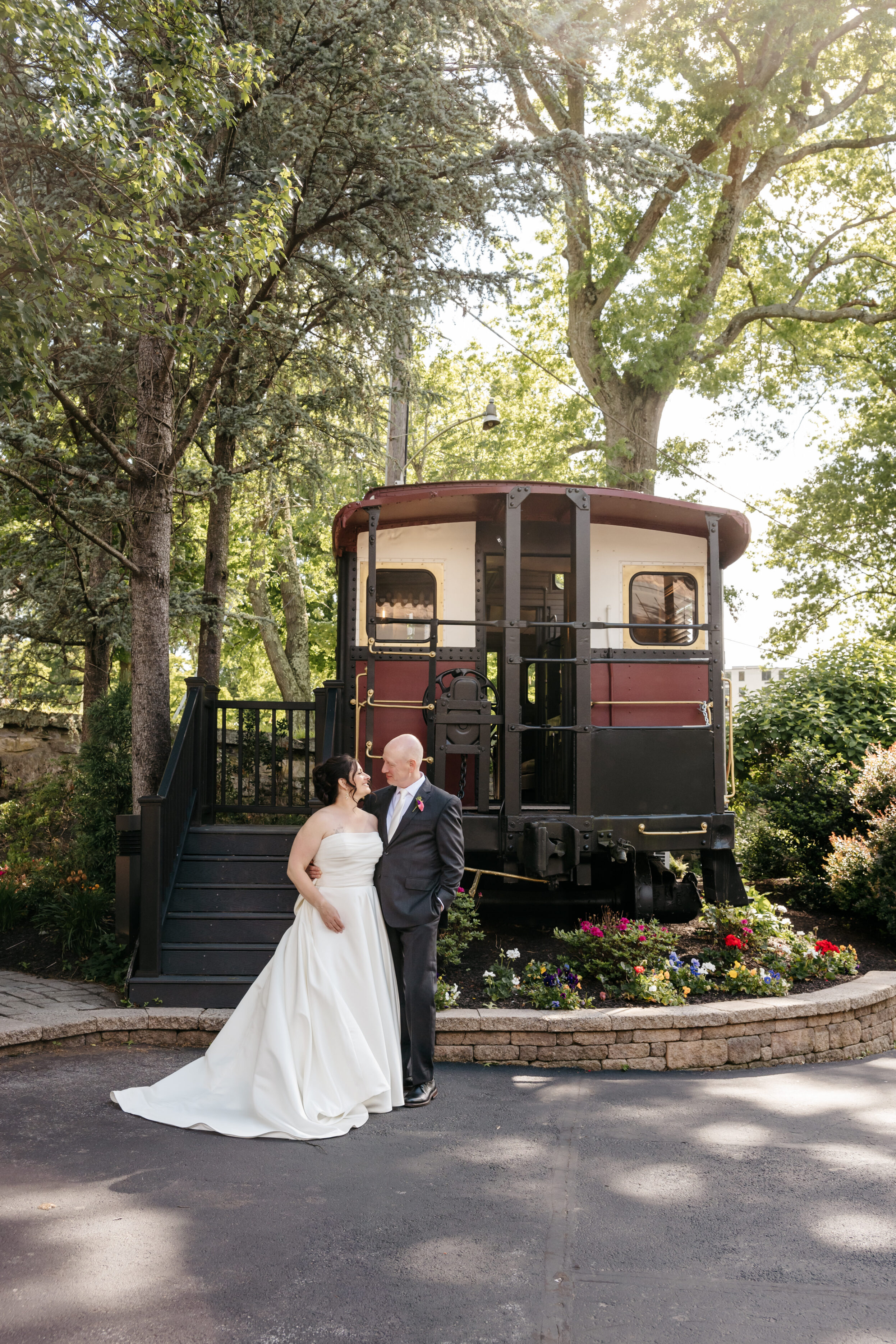 A bride and groom stand together, smiling, in front of a vintage red and cream train car surrounded by trees.