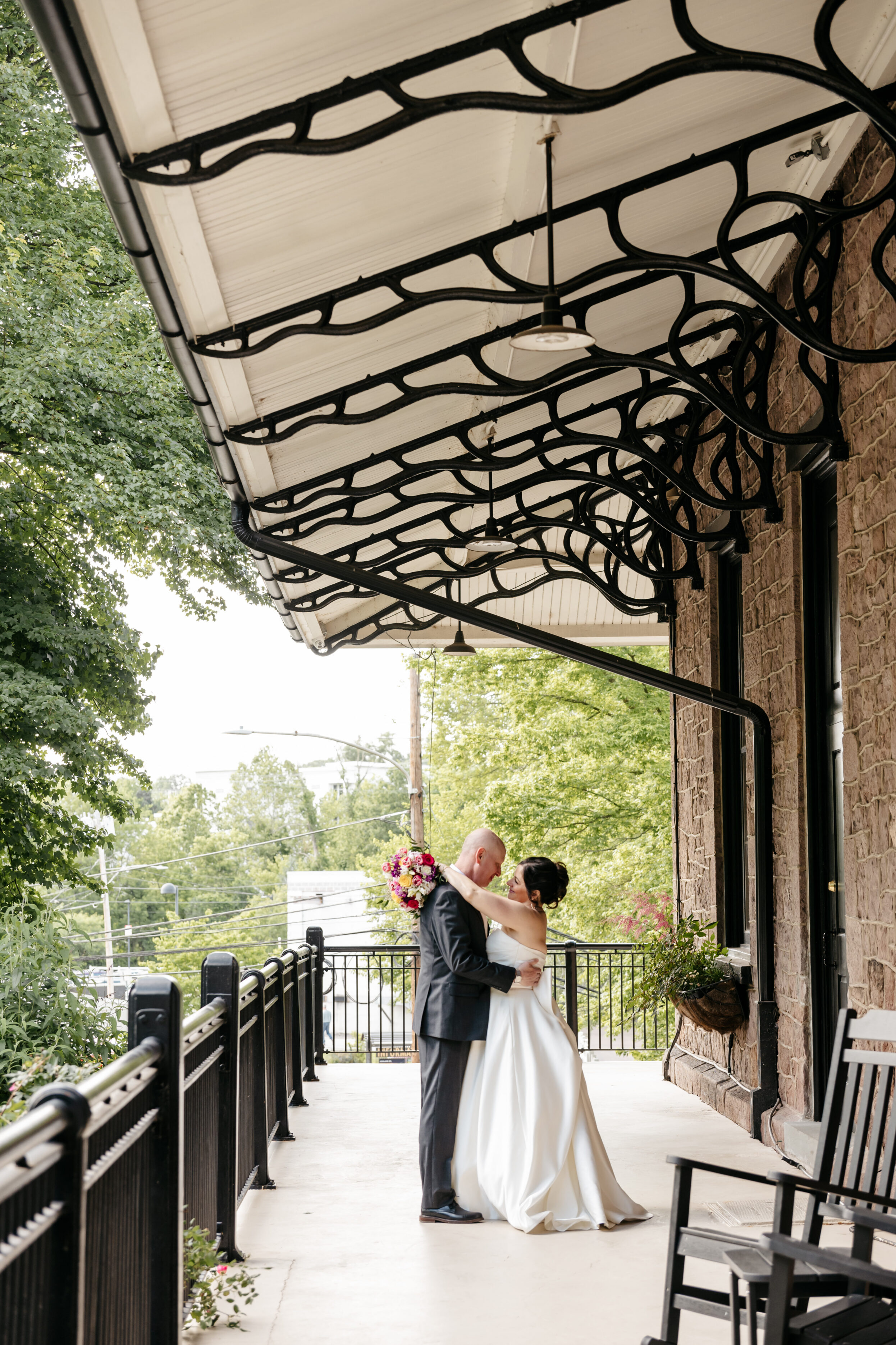 A bride and groom embrace on a covered outdoor walkway, surrounded by greenery and decorative iron railings.