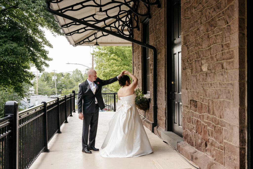 A bride and groom dance together on an outdoor walkway next to a stone building under a metal awning.