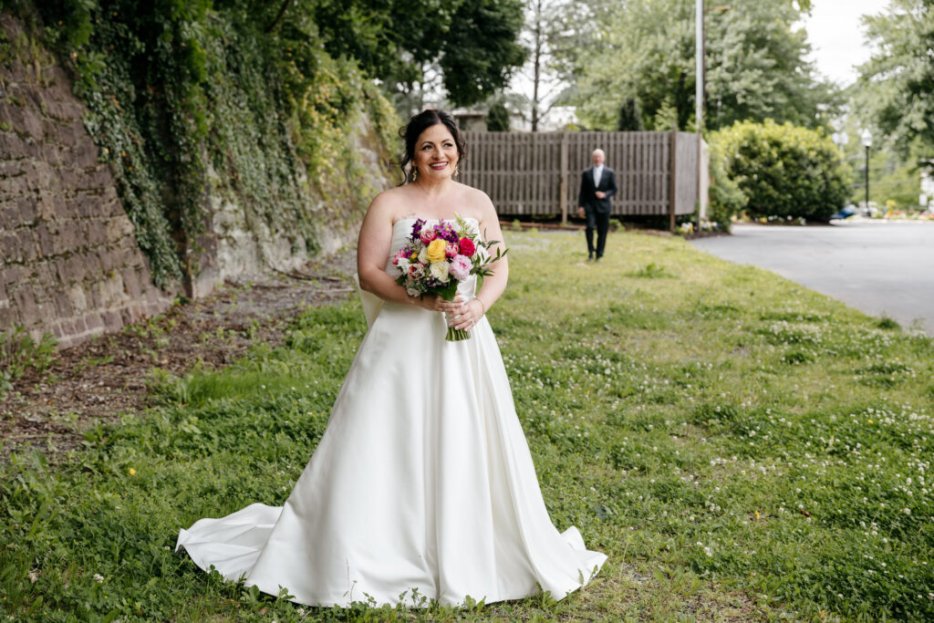 A bride in a white dress holds a bouquet and smiles outdoors, with a man walking in the background.