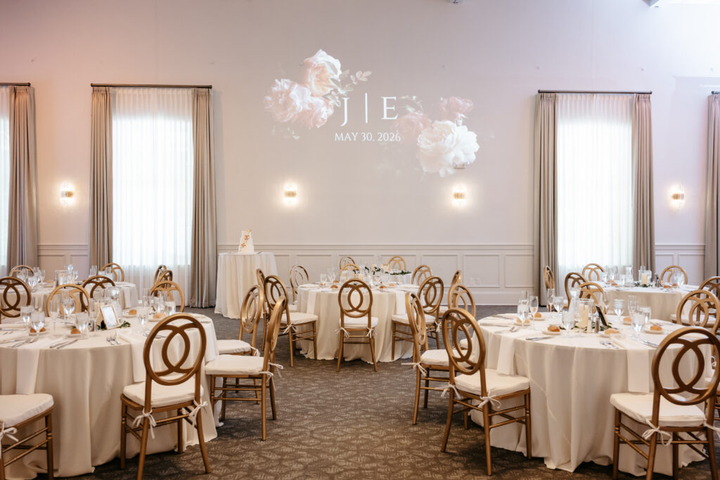 Elegant wedding reception room with round tables, gold chairs, and floral initials projected on the wall.