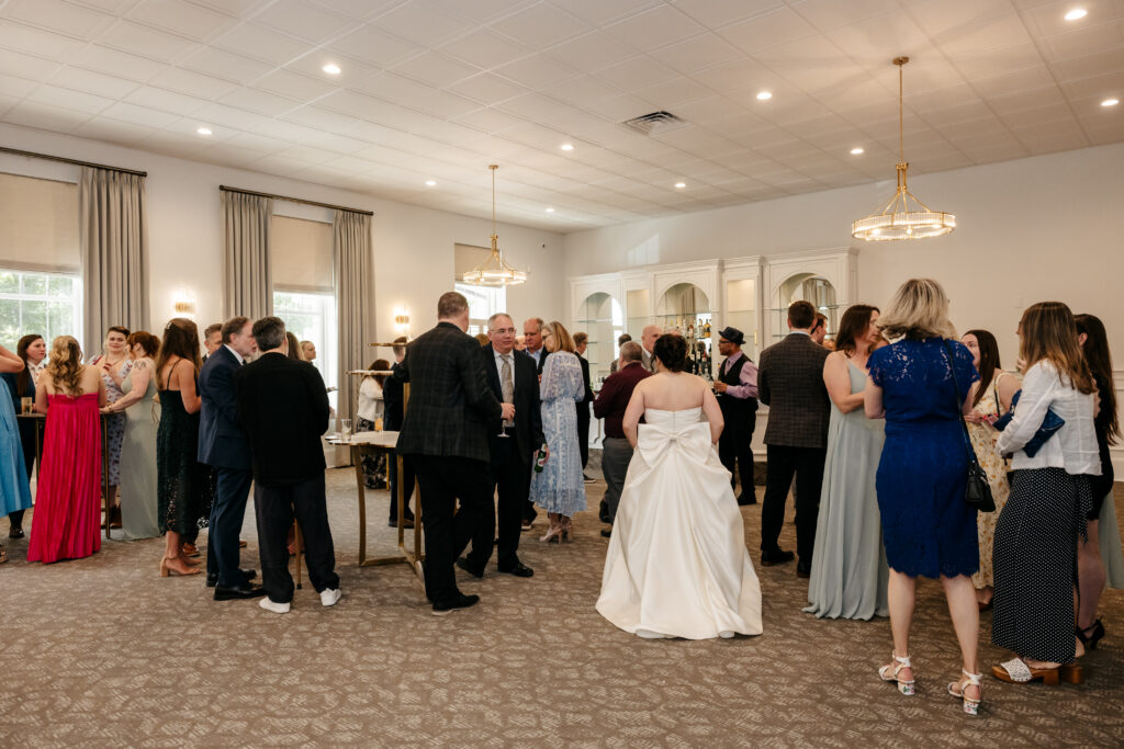 Wedding guests mingle in a bright reception room; a bride in a white dress stands in the center.