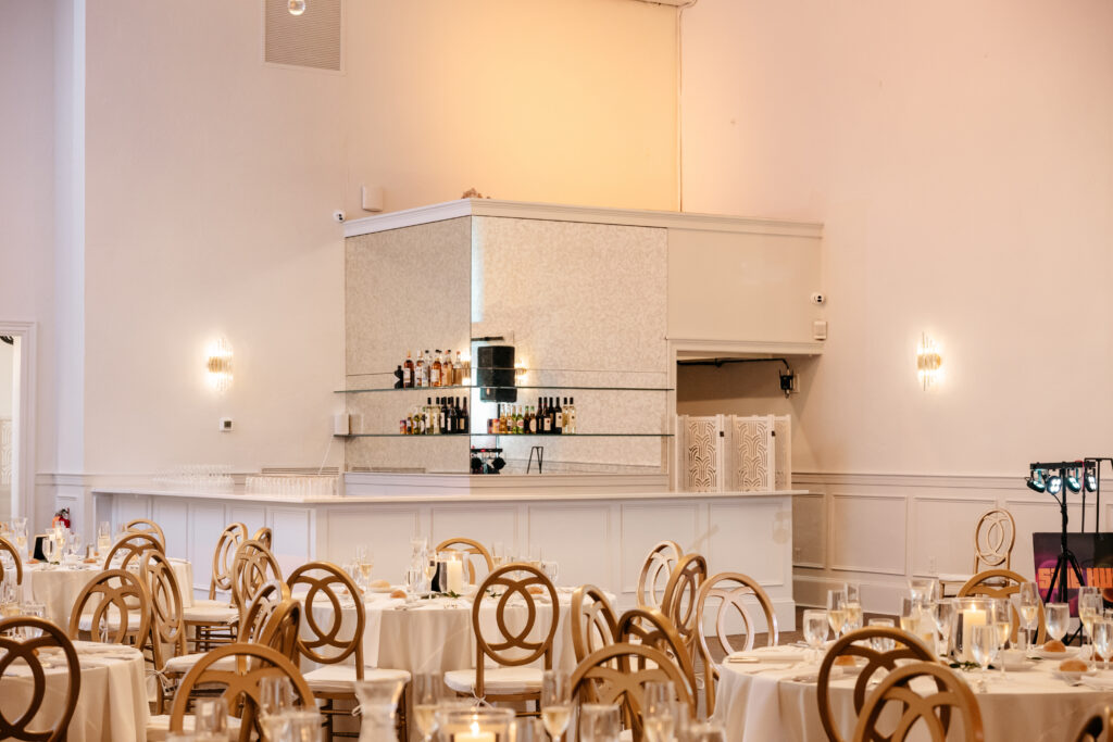 Elegant dining room with round tables, gold chairs, and a small wall-mounted bar with bottles and glassware.