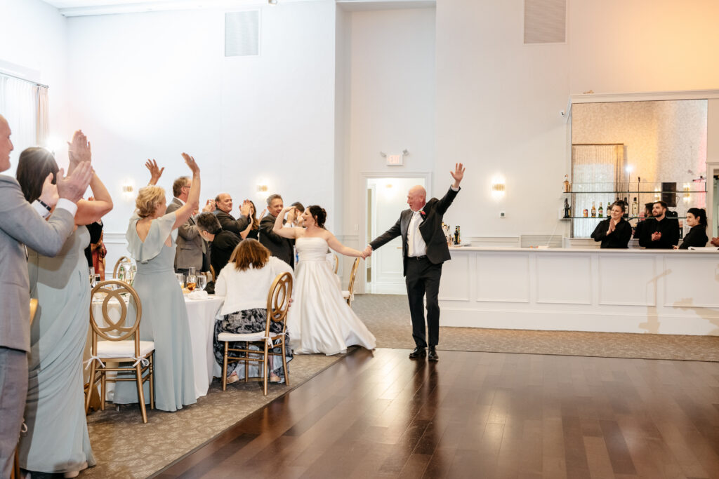A bride and groom enter a reception as guests cheer and clap, with a bar and staff visible in the background.