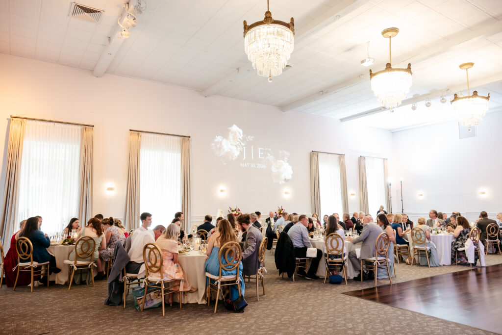 Guests seated at round tables in a bright, elegant banquet hall with chandeliers and tall windows.