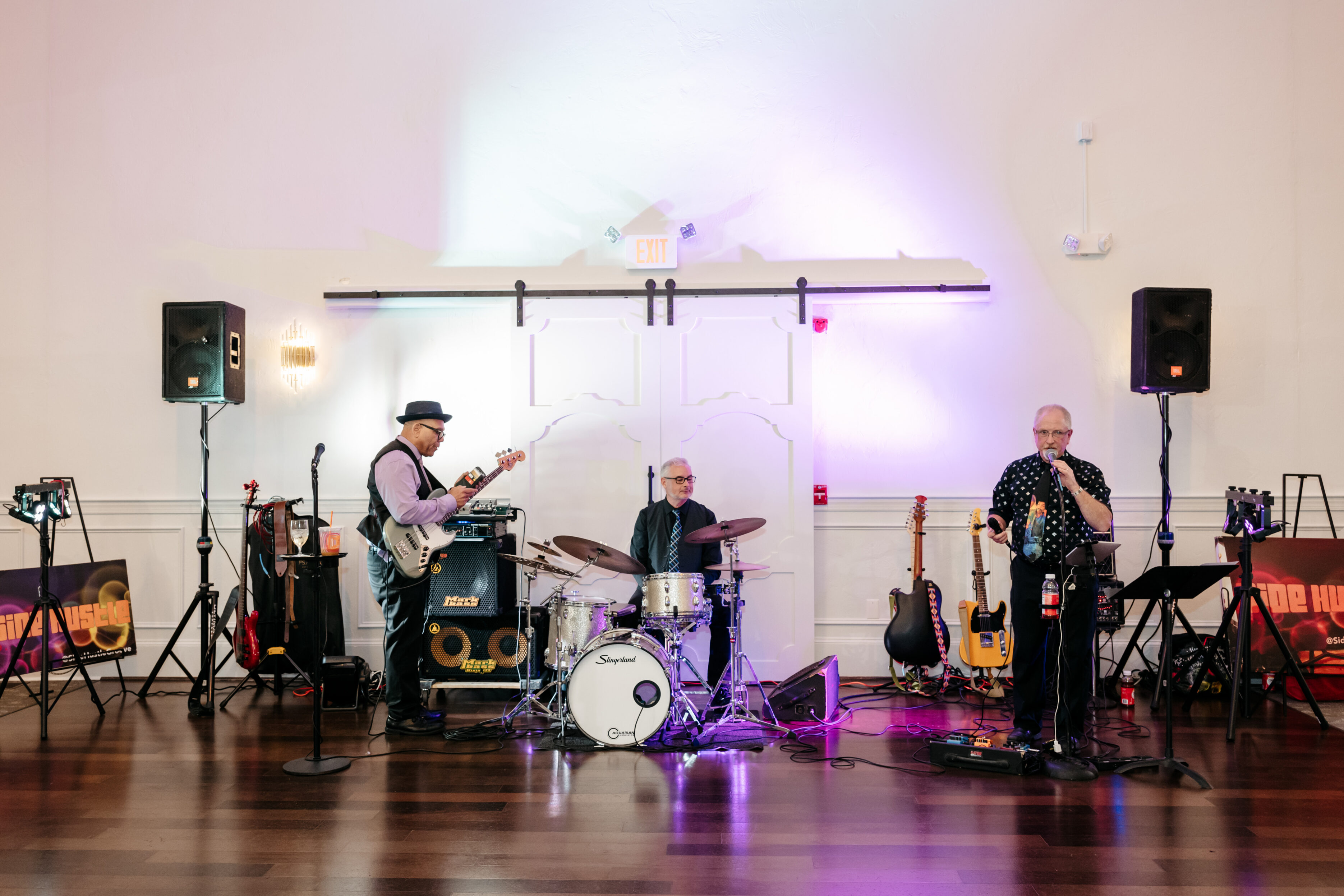 Three male musicians perform indoors on stage with guitars, drums, and microphone under purple lighting.