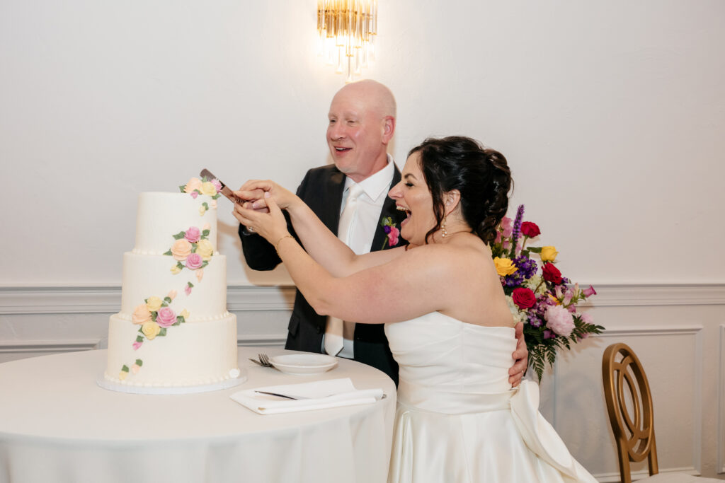A bride and groom joyfully cut their wedding cake together, smiling in a brightly lit reception room.
