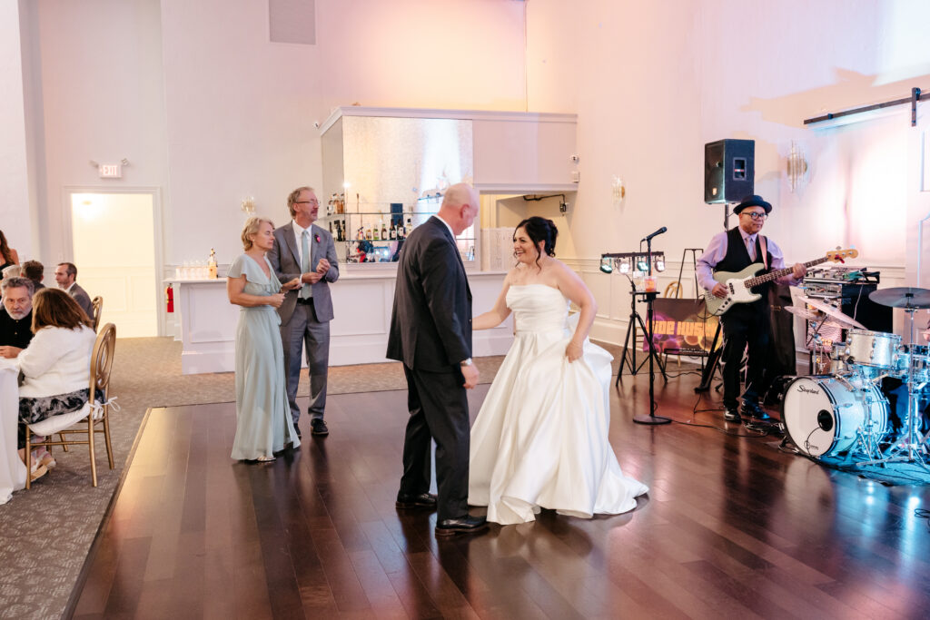 A bride in a white dress dances with an older man at a wedding reception while guests and a band look on.