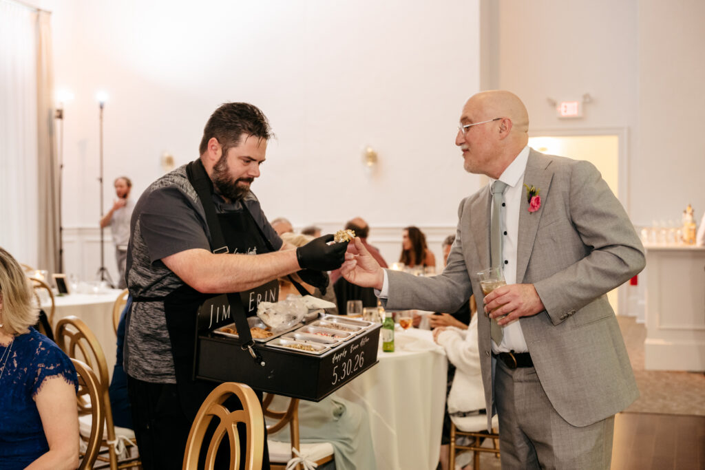 A server hands a small item to a guest in a gray suit at a formal indoor event with seated attendees.
