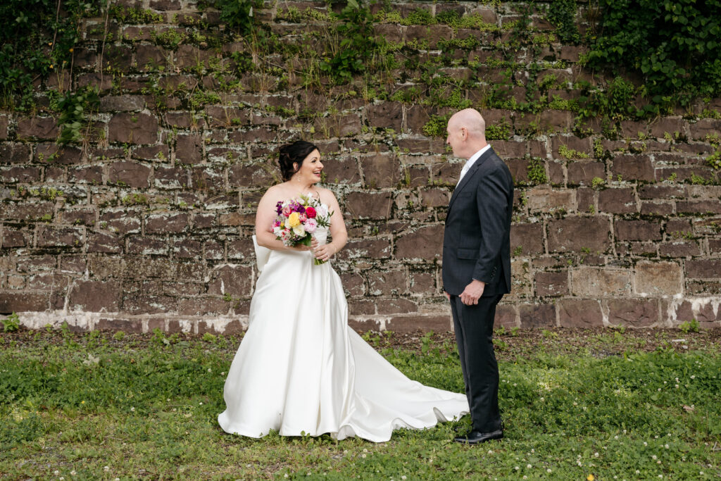 Bride in white dress holding bouquet smiles at groom in suit, standing on grass before a stone wall.