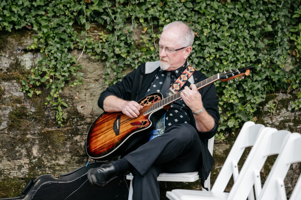 Man with glasses plays an acoustic guitar while seated outdoors beside empty white chairs and a wall covered in ivy.