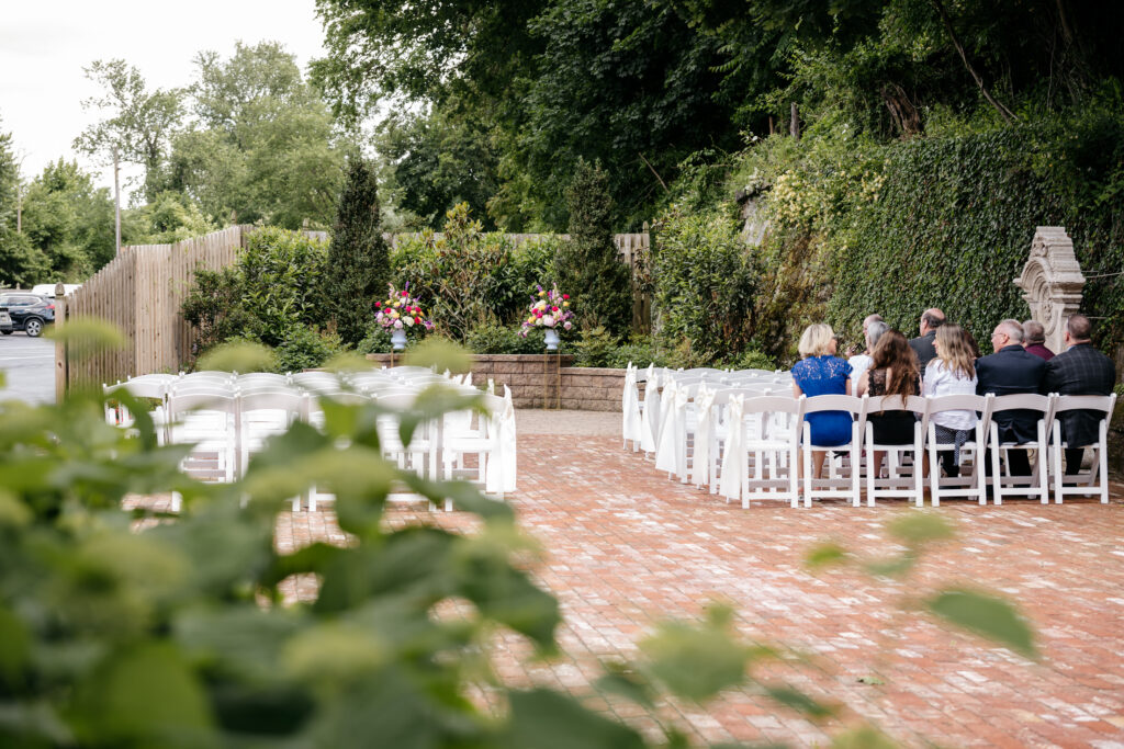 Outdoor wedding setup with white chairs, a few seated guests, and floral arrangements at the front altar.