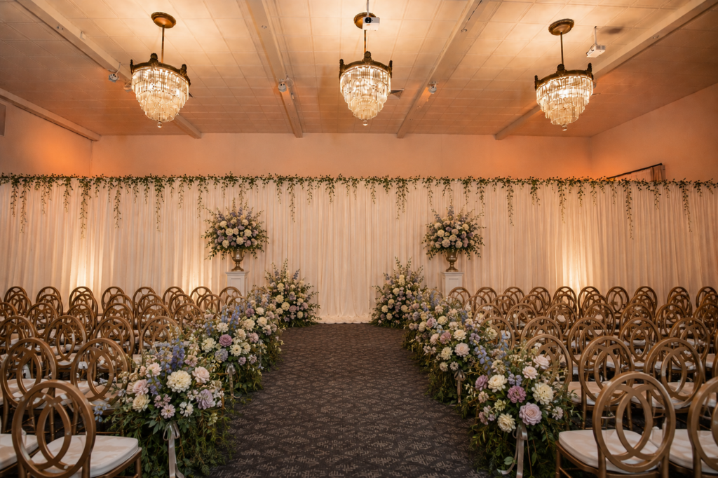 Elegant indoor wedding aisle with gold chairs, floral arrangements, chandeliers, and a draped backdrop.