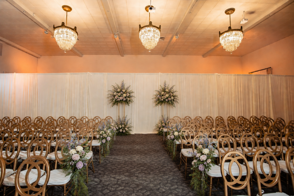 Indoor wedding ceremony setup with gold chairs, floral aisle decorations, and chandeliers overhead.