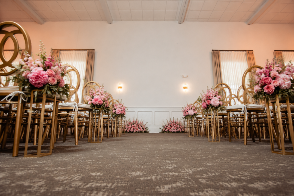Rows of gold chairs with pink flower arrangements line an aisle in an elegant, softly lit indoor venue.