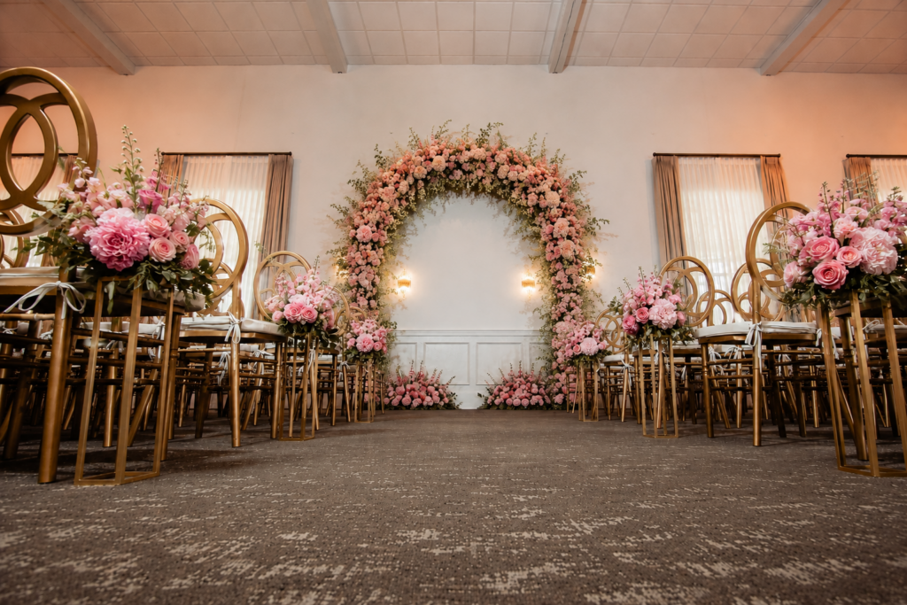 Indoor wedding aisle with gold chairs and pink floral arrangements, facing a large, flower-decorated arch.