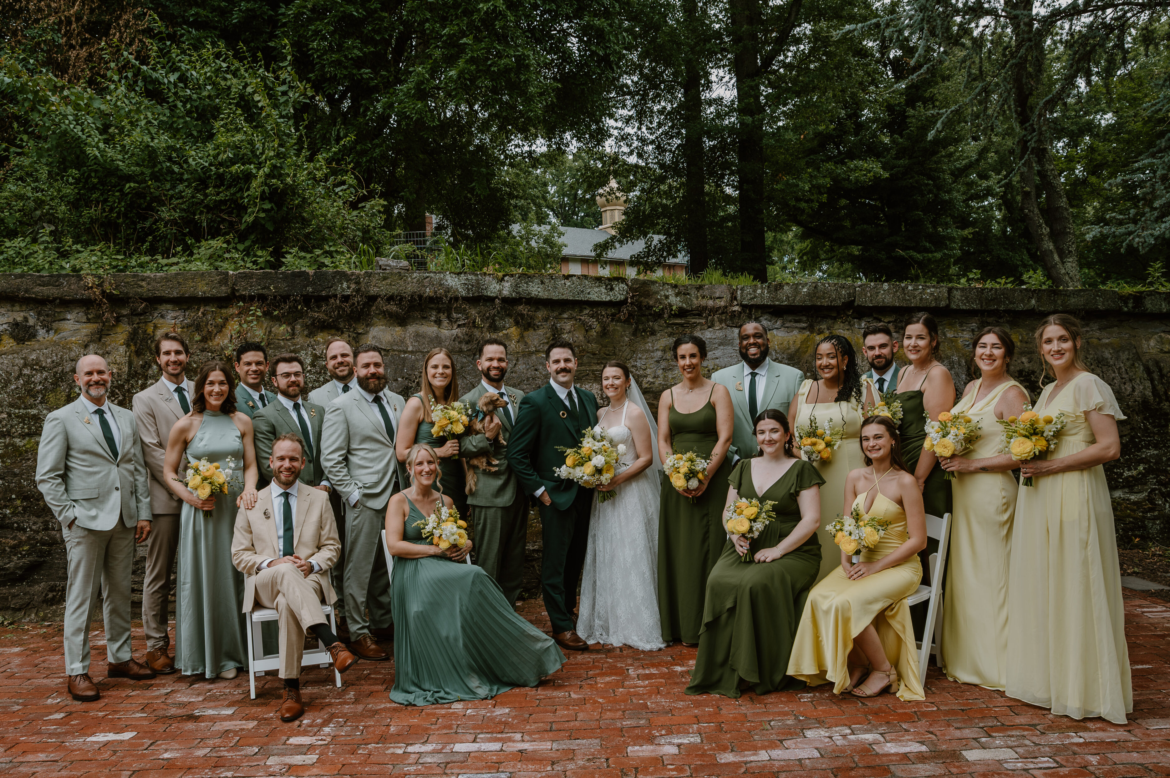 Wedding party posing outdoors; bride and groom center, surrounded by bridesmaids and groomsmen in green and yellow attire.