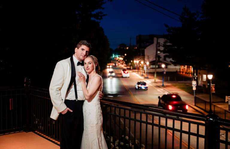 Bride and groom embrace on a balcony at night, overlooking a street with cars and glowing streetlights.