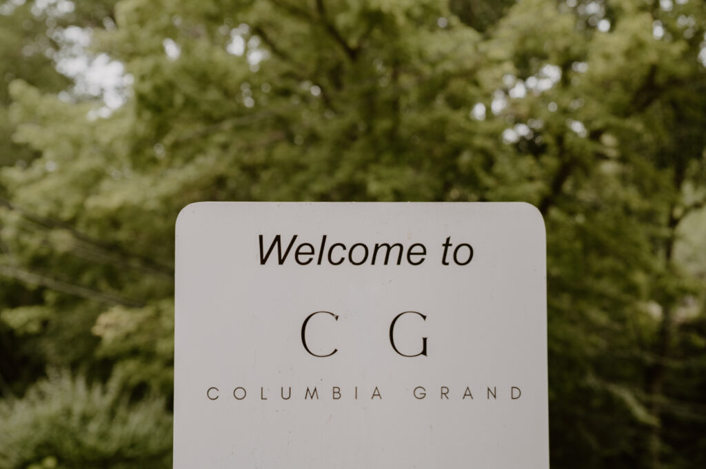 A sign reads “Welcome to C G Columbia Grand” with trees and greenery blurred in the background.