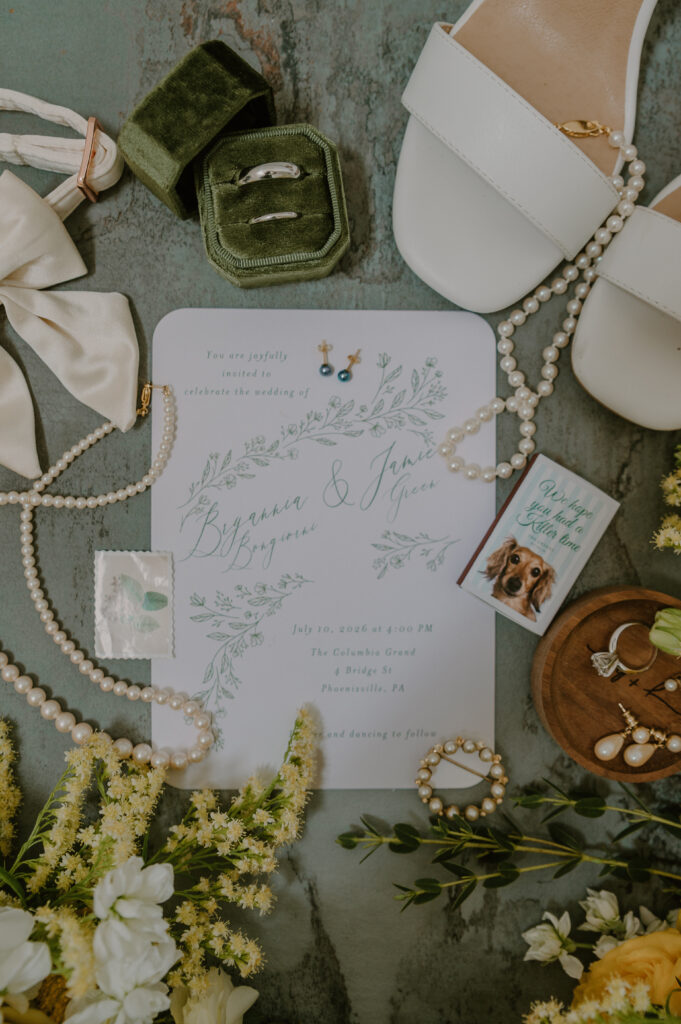 Wedding invitation flat lay with jewelry, shoes, flowers, a ring box, and a small dog-themed matchbox.