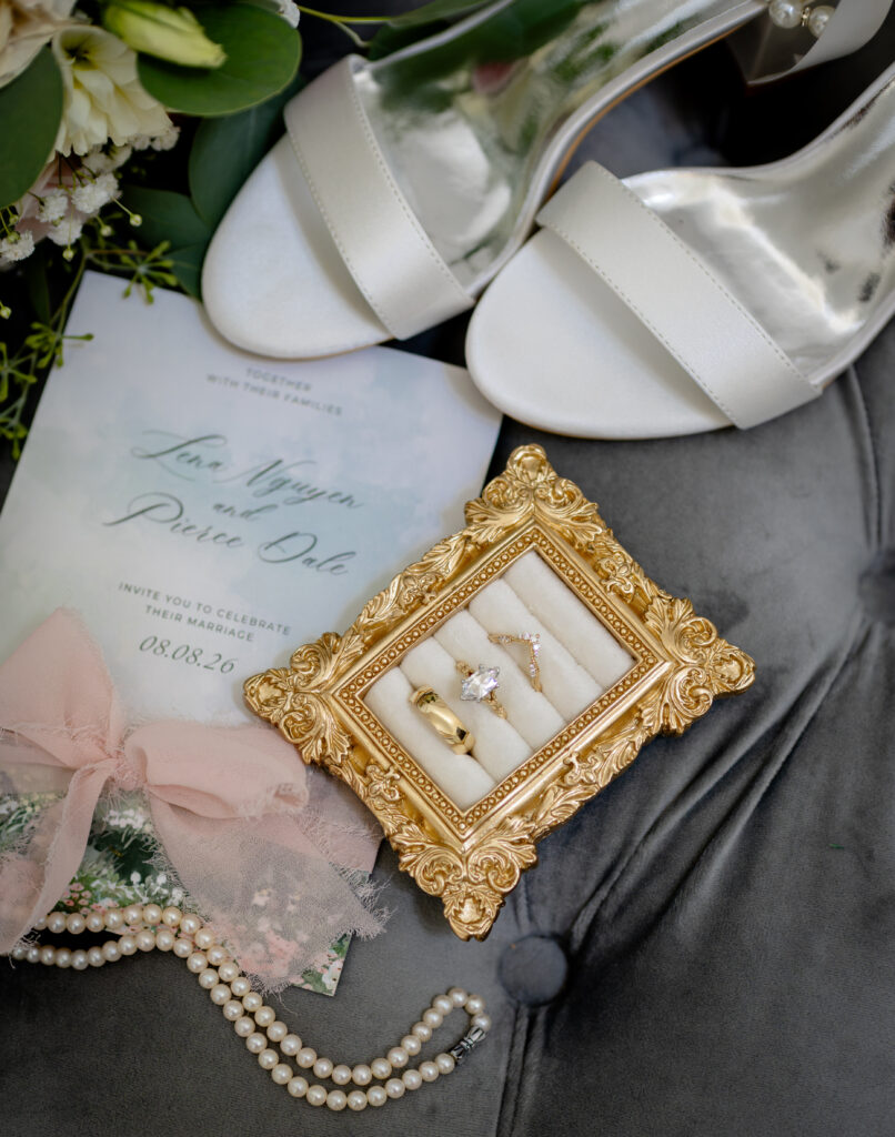 Wedding invitation, white heels, pearl bracelet, and gold-framed rings on a gray surface with flowers.
