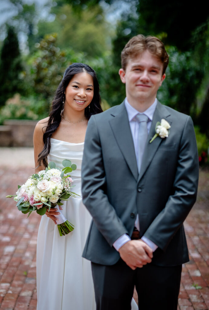 A bride holding a bouquet smiles behind a groom in a suit outdoors on a brick path.
