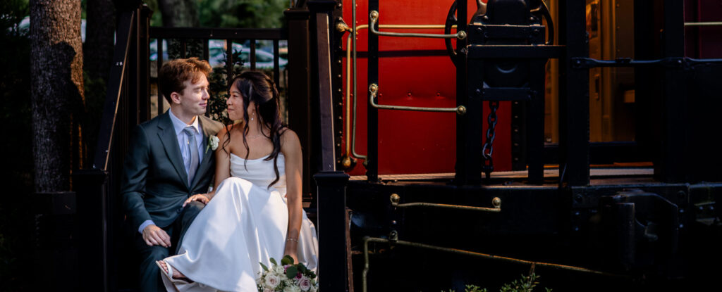 A bride and groom sit on steps by a vintage red train car, surrounded by greenery in the evening light.