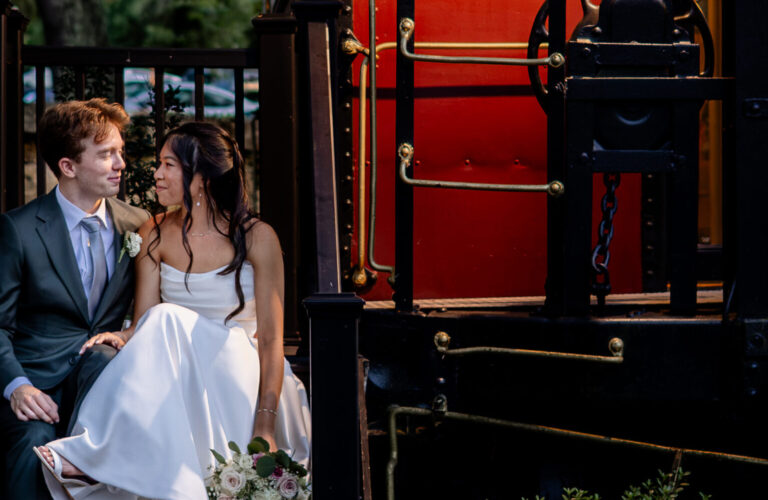 A bride and groom sit on steps by a vintage red train car, surrounded by greenery in the evening light.