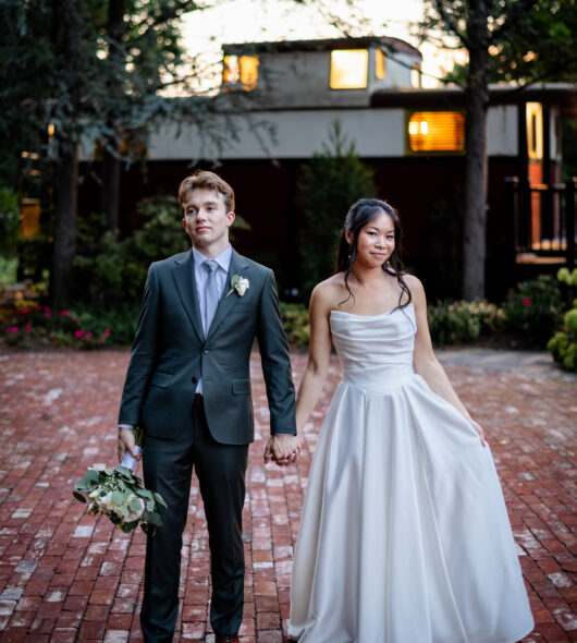Bride in white dress and groom in suit holding hands outdoors on brick path, greenery and house in background.