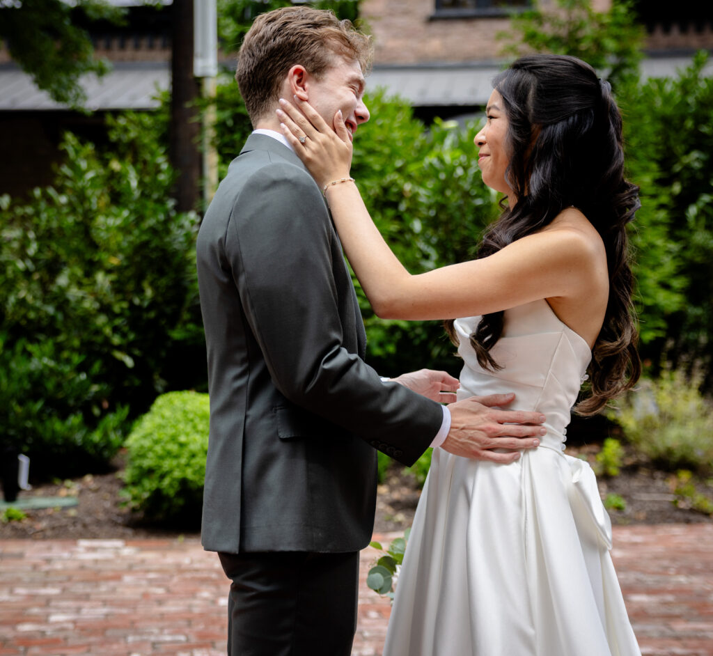 A bride in a white gown smiles and touches the groom’s face outdoors, both looking happy.