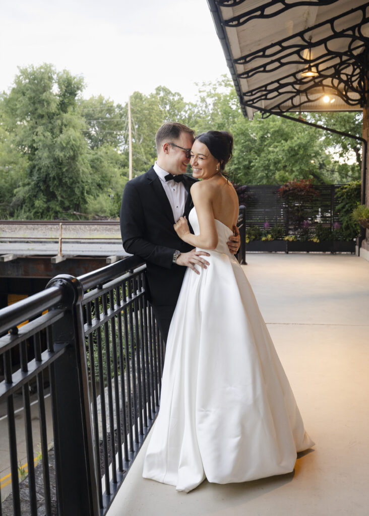 A bride and groom smile together on a covered outdoor terrace, surrounded by greenery.